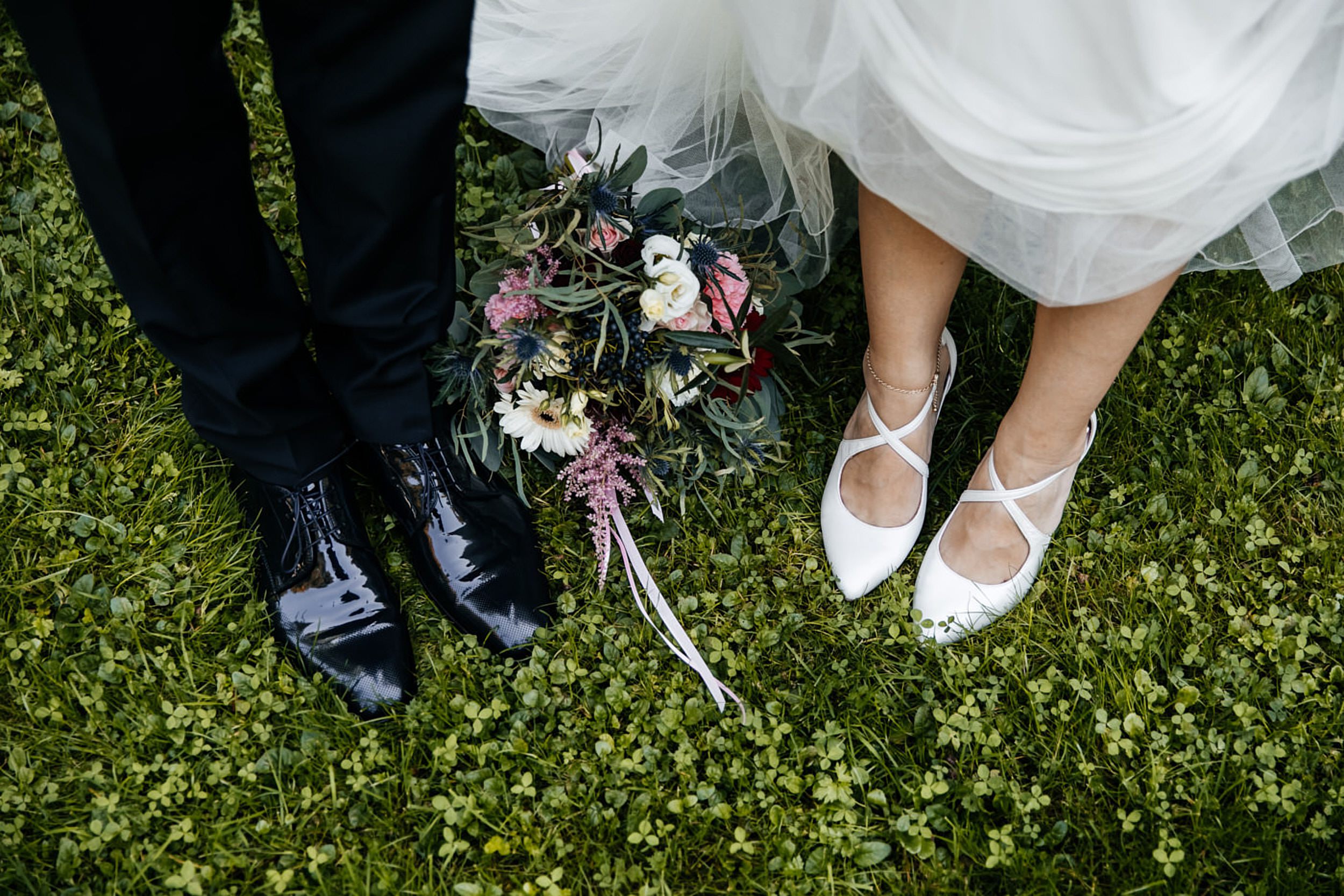 Brautpaar-Shooting, Detailaufnahme. Schuhe von Bräutigam der Braut neben dem Braut-Strauß auf dem Rasen. Begleitung als Hochzeitsfotograf in Detmold.