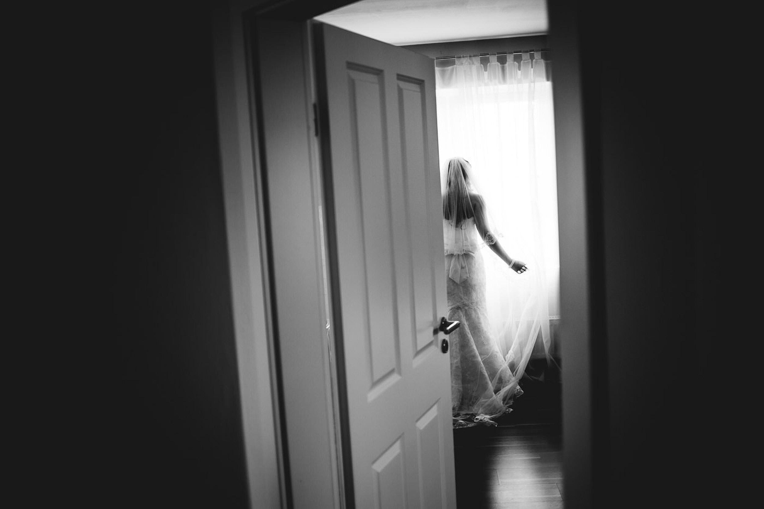 Halb geöffnete Tür. Die Braut steht am Fenster und spielt mit dem Schleier.