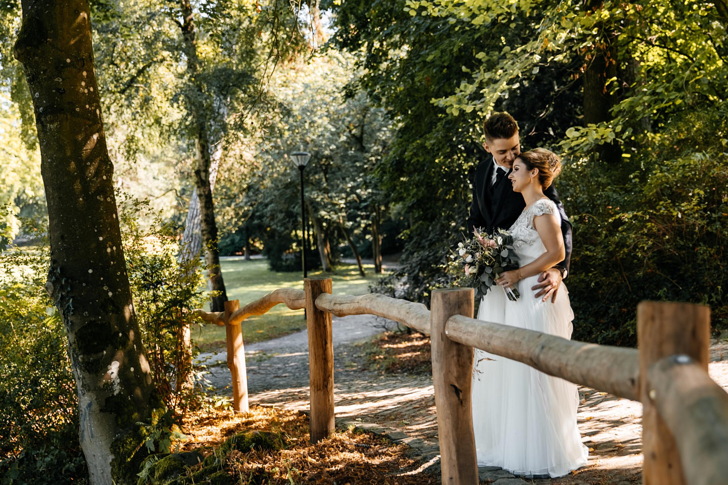 Fotoshooting am Hochzeitstag. Pala Garten in Detmold Brautpaar steht an dem Holzzaun der Bräutigam küsst die Braut auf den Kopf. Hochzeitsfotograf in Detmold.