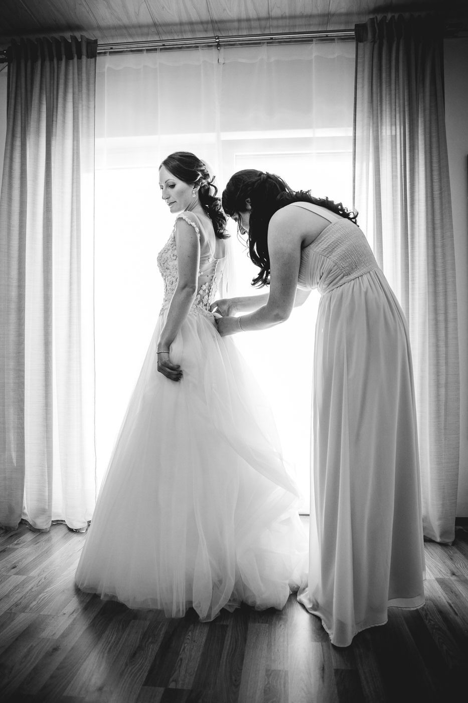 Trauzeugin macht der Braut am lichterfülltem Fenster das Kleid zu, schwarz weiß. Hochzeitsfotograf in Bad Ems.