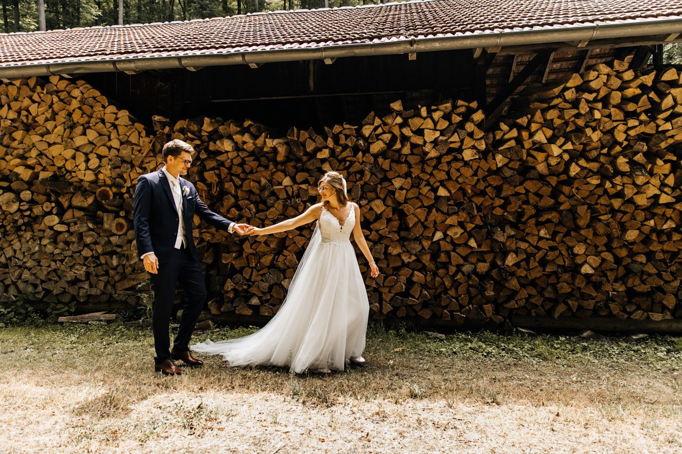Fotoshooting am Hochzeitstag auf dem Bauernhof. Brautpaar geht an dem Holzunterstand Hand-in-Hand. 